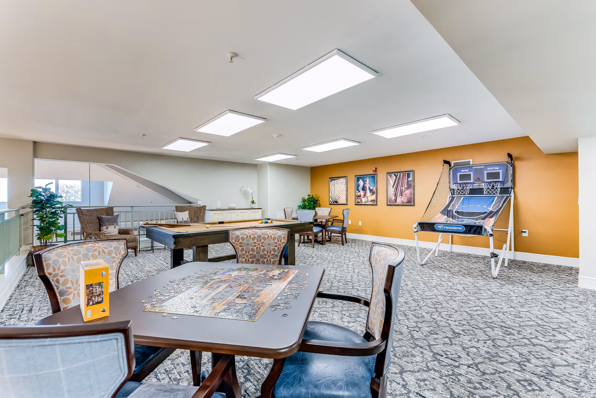 A bright and spacious recreational room in a senior living facility featuring a table with a partially completed jigsaw puzzle, several chairs with patterned upholstery, a shuffleboard table, and an electronic basketball shooting game against a mustard-colored wall with framed artwork. The room has carpeted flooring, large ceiling lights, and some potted plants near the windows.