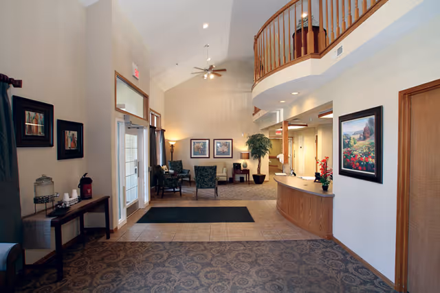 Interior view of a senior living facility lobby with a reception desk on the right, seating area with chairs and tables in the background, decorative paintings on the walls, a potted plant, and a high ceiling with a ceiling fan.