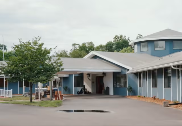 Exterior view of a single-story assisted living facility building with a covered entrance, blue walls, and multiple windows. There is a tree and some benches near the entrance, and the sky is cloudy.