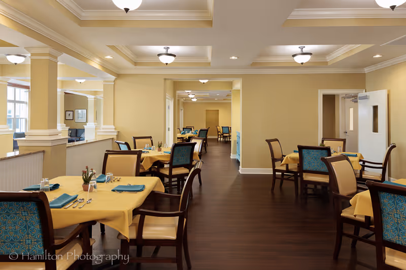 A well-lit dining room with multiple tables covered in yellow tablecloths, each set with blue napkins, silverware, glasses, and small potted plants. The room features beige walls, dark wood flooring, and ceiling lights with white shades. Chairs have wooden frames with beige and blue patterned upholstery. The space appears clean and organized, designed for communal dining.