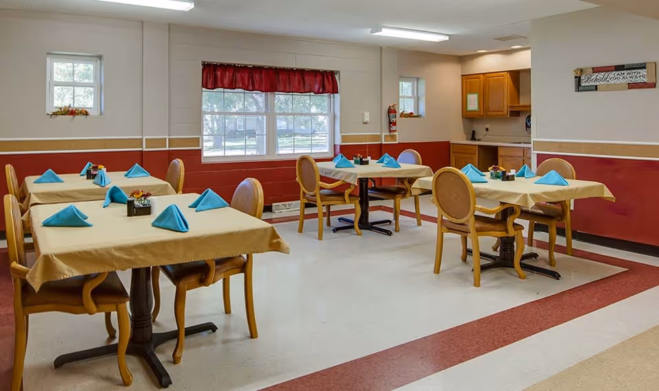 A dining room with four tables covered in beige tablecloths, each set with blue folded napkins and small flower arrangements. The room has red and beige walls, three windows with white frames, and wooden chairs with cushioned seats. There is a small kitchenette area with wooden cabinets in the background.