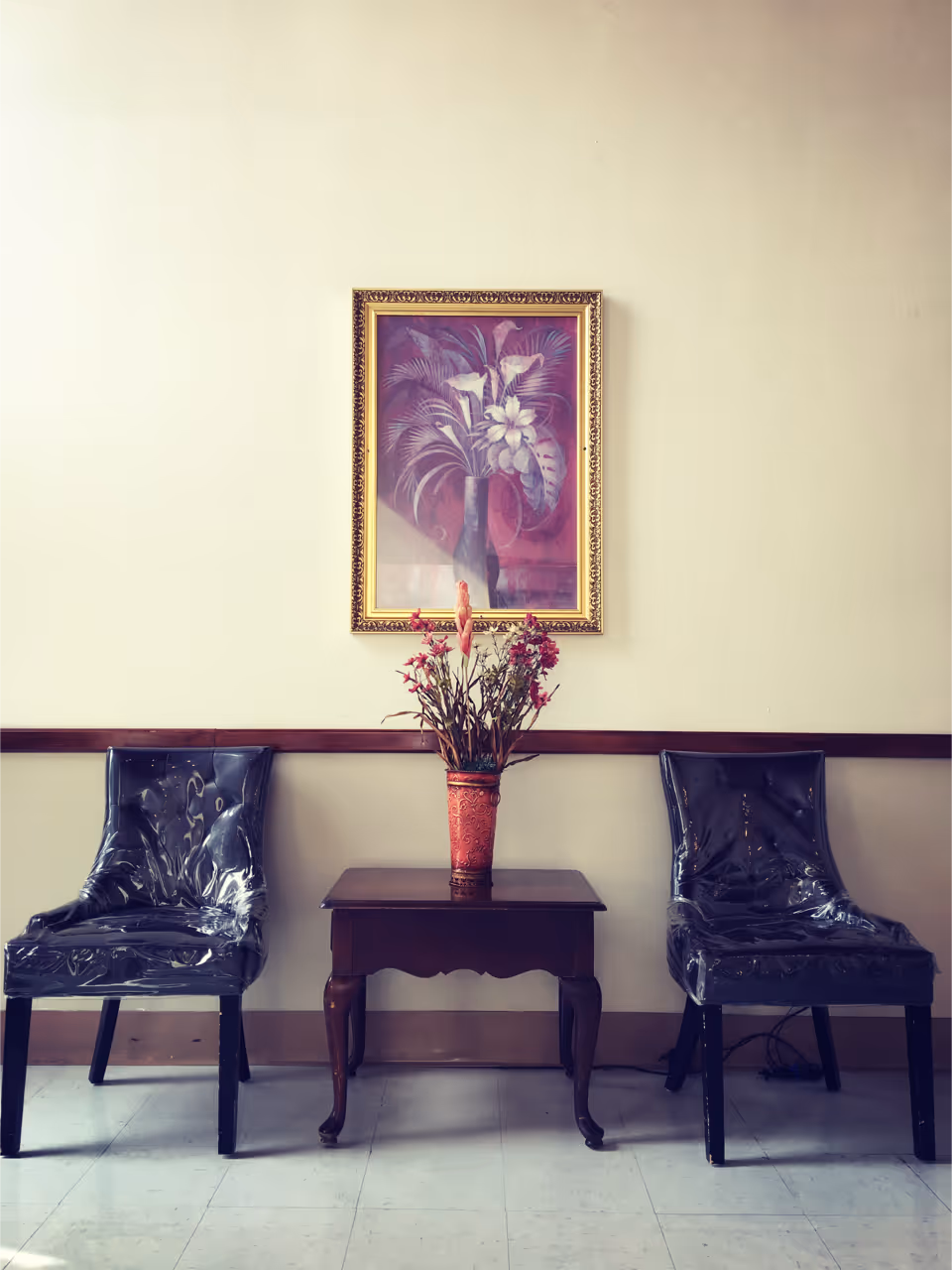 Two black chairs covered in plastic are placed on either side of a small wooden table with a red vase containing flowers. Above the table, a framed painting of a floral arrangement hangs on a beige wall.