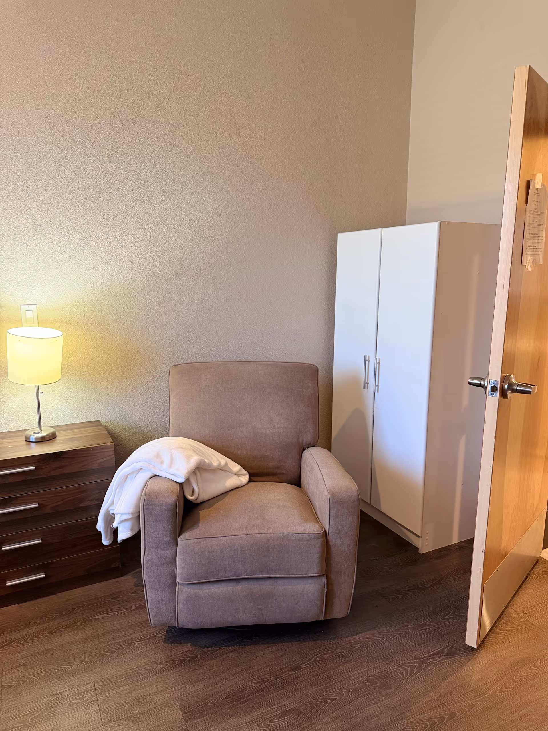 A cozy corner of a room featuring a beige upholstered armchair with a white blanket draped over one arm. To the left of the chair is a wooden nightstand with three drawers and a lit table lamp on top. To the right, there is a white wardrobe with double doors. The room has wooden flooring and a partially open wooden door on the right side.