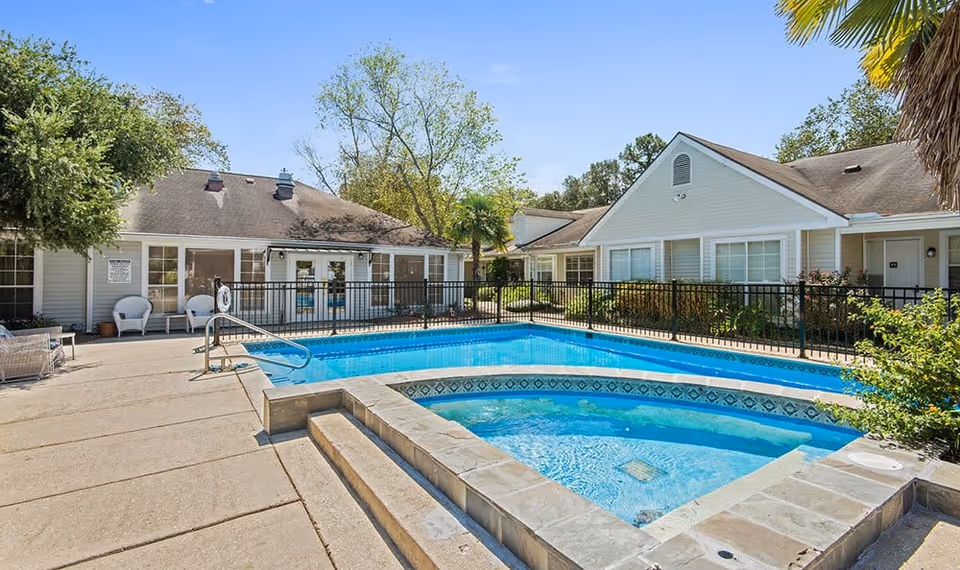 Outdoor swimming pool and hot tub area at Southside Gardens facility, surrounded by a concrete deck, black metal fence, and single-story residential-style buildings with large windows and trees in the background under a clear blue sky.