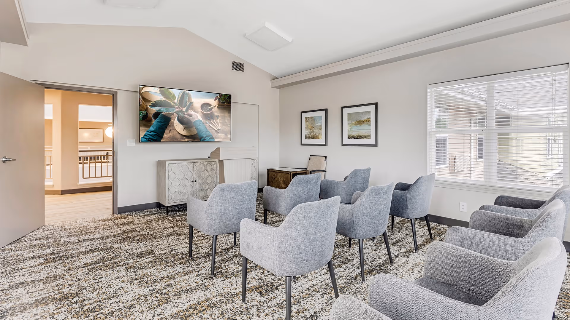 A bright and modern common area with eight gray upholstered chairs arranged in two rows facing a wall-mounted flat screen TV. The room has a patterned carpet, light-colored walls, two framed landscape paintings, a window with blinds, and an open door leading to a hallway.