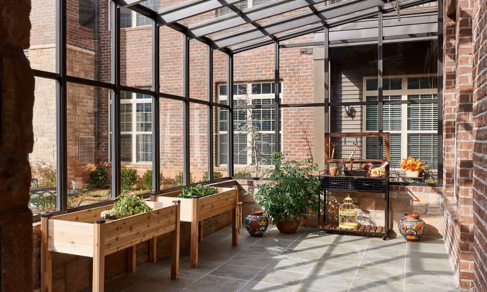 Sunlit indoor garden area with large glass windows and a glass ceiling. There are two wooden planter boxes with green plants, a potted leafy plant, colorful decorative pots, and a black metal cart with a yellow birdcage and other decorative items. The walls are made of brick and stone.