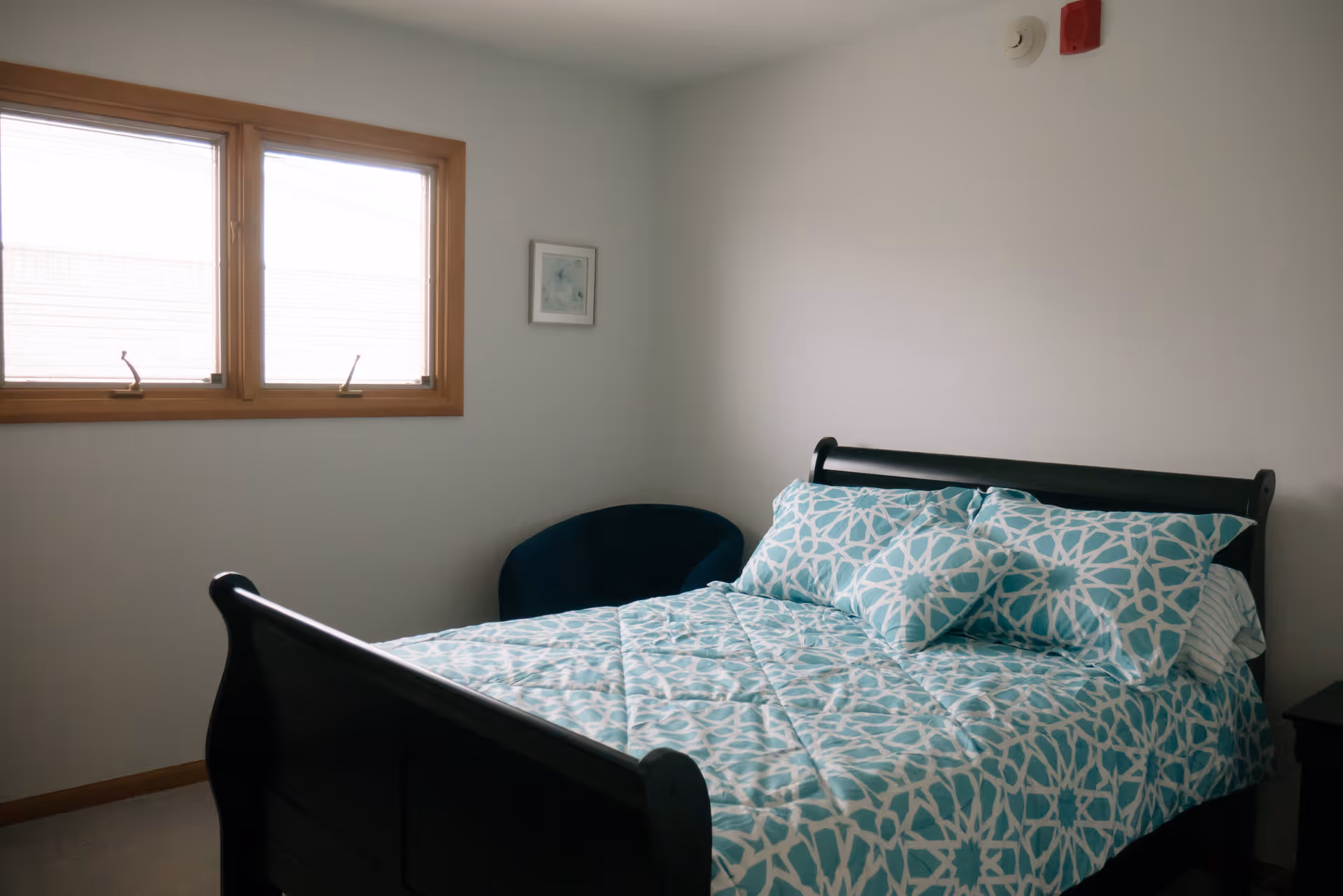 A bedroom with a dark wooden bed frame, a bed made with a blue and white patterned comforter and matching pillows, a dark blue chair in the corner, two windows with wooden frames, and a small framed picture on the wall.