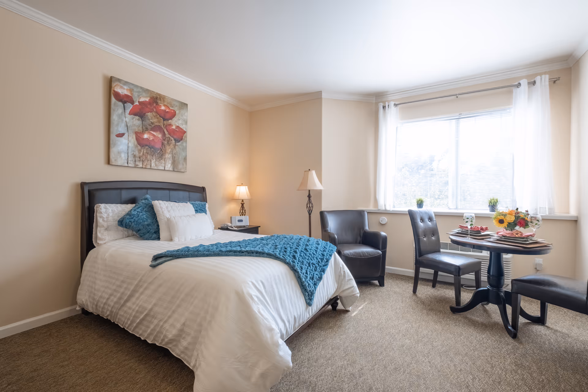 A cozy bedroom with a neatly made bed featuring white linens and blue accent pillows and throw. There is a bedside table with a lamp and a clock, a floor lamp, a small round table set for two with plates and glasses, and two black chairs near a large window with white curtains. A painting of red flowers hangs above the bed.