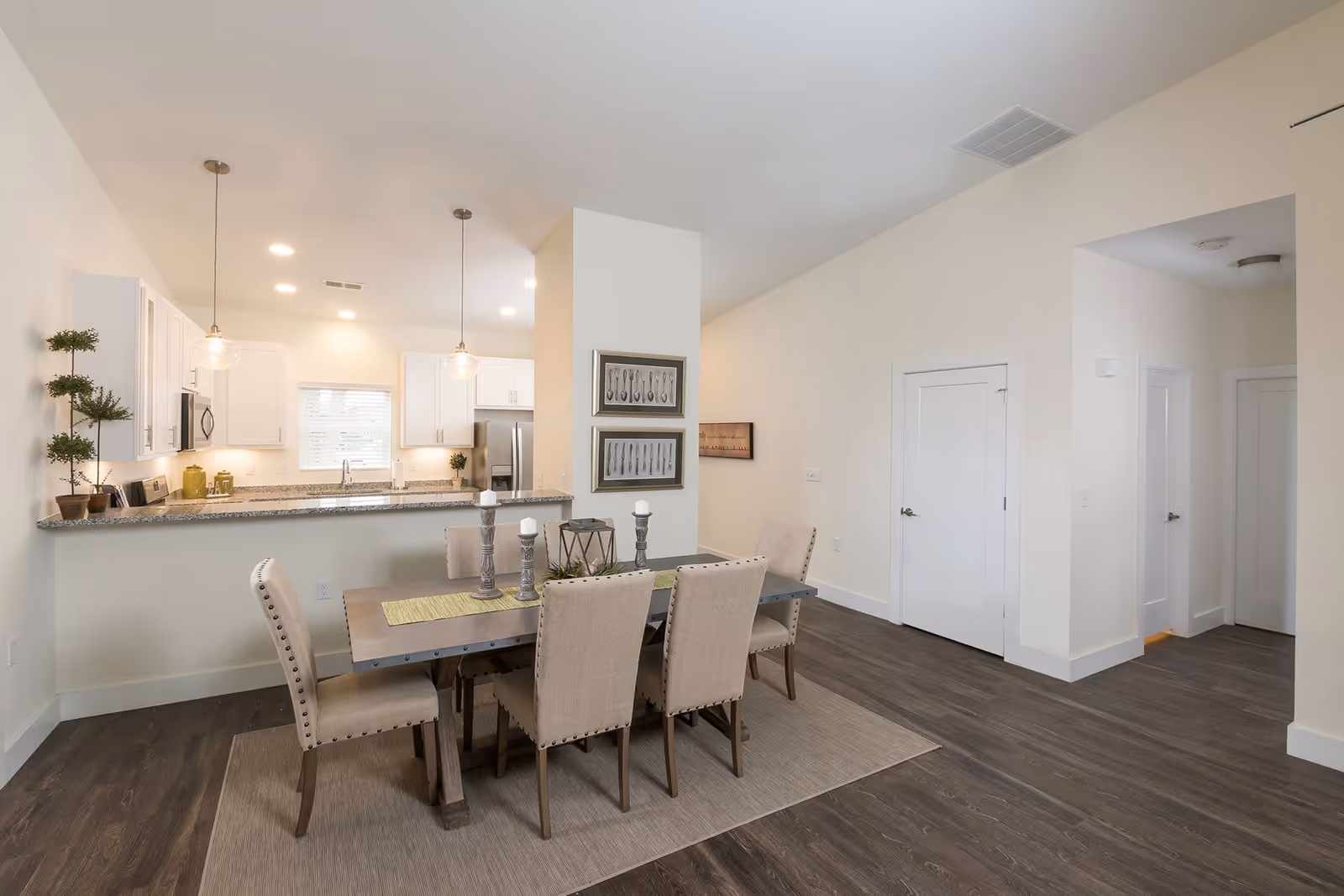 A modern dining area with a rectangular table surrounded by six beige upholstered chairs on a light-colored rug. The dining area is adjacent to an open kitchen with white cabinets, granite countertops, stainless steel appliances, and two pendant lights hanging over the counter. The room has light-colored walls and dark wood flooring.