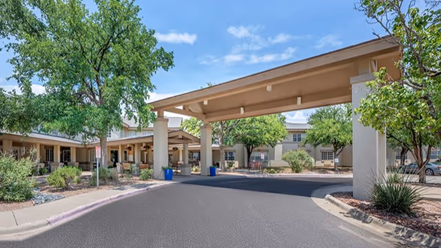 Entrance area of a senior living facility with a covered driveway, surrounded by trees and shrubs, under a partly cloudy blue sky.