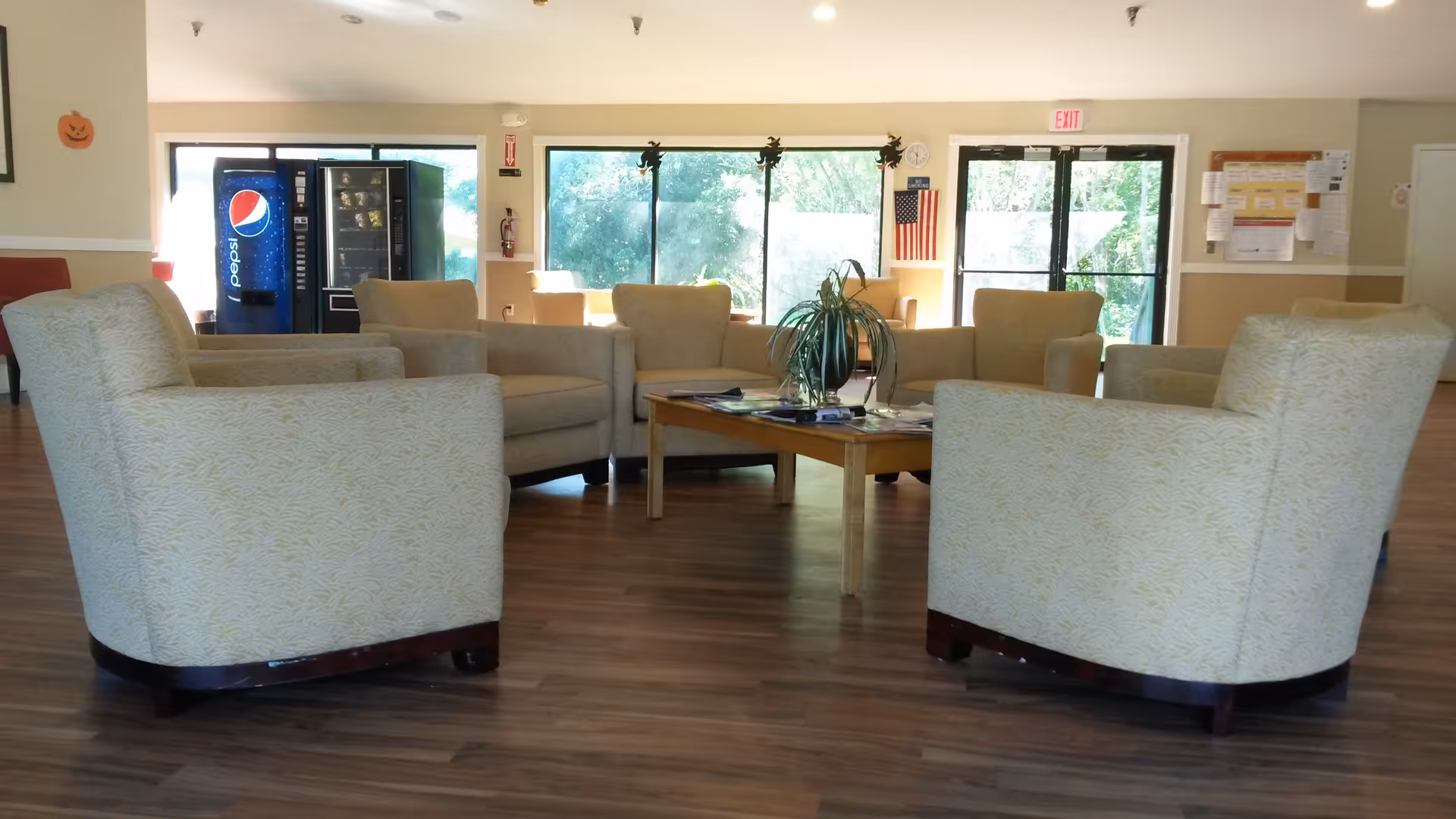 A common area with several beige upholstered armchairs arranged around a wooden coffee table with a plant and magazines on it. Large windows and glass doors let in natural light, and a Pepsi vending machine is visible in the background. The room has wood flooring and light-colored walls with some decorations, including small Halloween-themed cutouts on the windows.