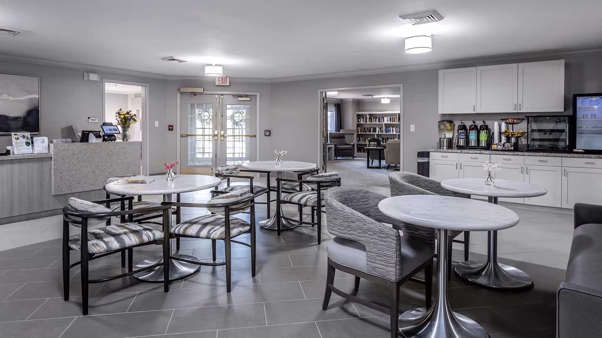 A modern common area in a senior living facility with round tables and cushioned chairs arranged on a tiled floor. There is a counter with coffee and beverage dispensers on the right side, and a reception desk with flowers on the left. Double glass doors and an open doorway leading to a room with bookshelves and armchairs are visible in the background.