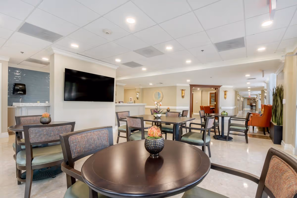 A bright and spacious common area in a senior living facility featuring several round tables with chairs arranged around them. Each table has a small decorative plant. A large flat-screen TV is mounted on a wall, and there are comfortable seating areas with armchairs along the hallway. The space has a clean, modern design with recessed lighting and light-colored walls and flooring.