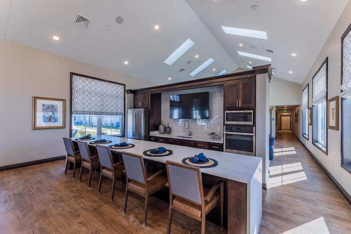 A modern kitchen and dining area with a long white island countertop set with six place settings, each with a blue napkin. The kitchen features dark wood cabinetry, a stainless steel refrigerator, built-in microwave and oven, a sink, and a large flat-screen TV mounted on a white tiled backsplash. The room has high ceilings with skylights and recessed lighting, and windows with patterned blinds letting in natural light. A hallway with windows and framed pictures is visible to the right.