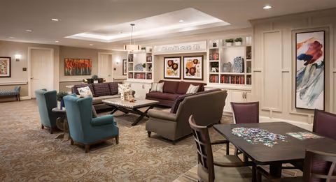 Communal lounge with sofas, armchairs, built-in bookshelves, artwork, and activity tables.