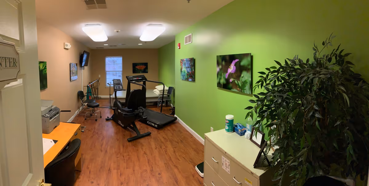 A small therapy or exercise room with wood flooring and walls painted green and beige. The room contains a treadmill, a stationary bike, a therapy bed, a desk with a chair, a filing cabinet, and a large potted plant. There are framed pictures of flowers on the walls and a window at the far end letting in natural light.