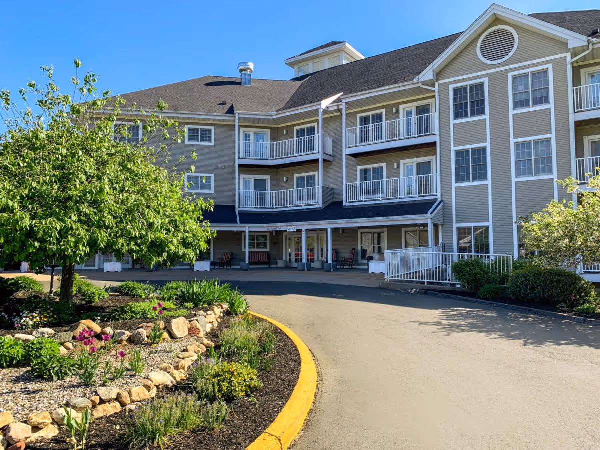 Exterior view of a multi-story senior living facility building with balconies, a covered entrance, and landscaped garden with flowers and shrubs in front under a clear blue sky.