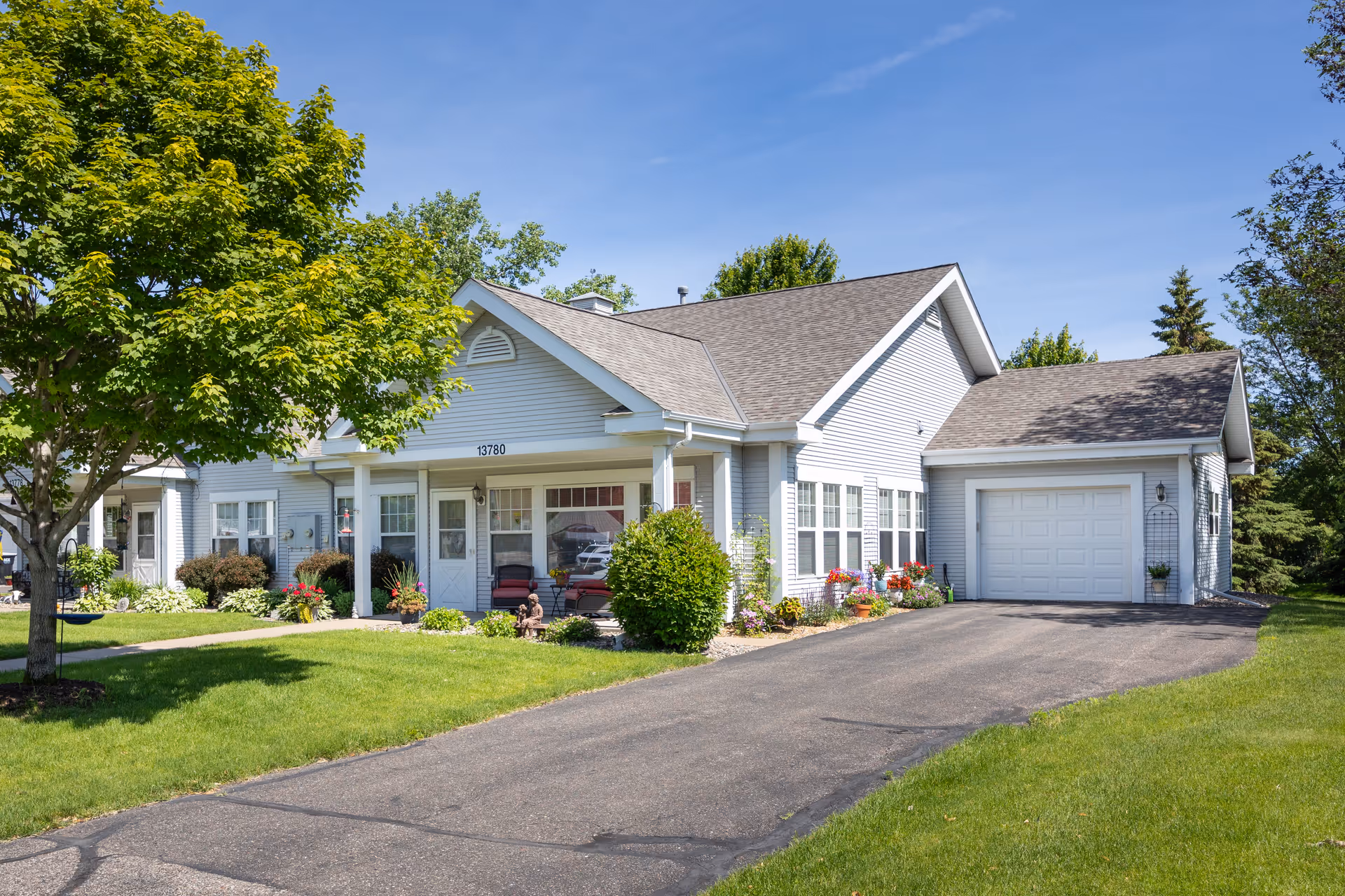 A single-story white house with a gray shingled roof, a front porch with chairs, a driveway leading to a garage, and a well-maintained lawn with trees and flower beds under a clear blue sky.