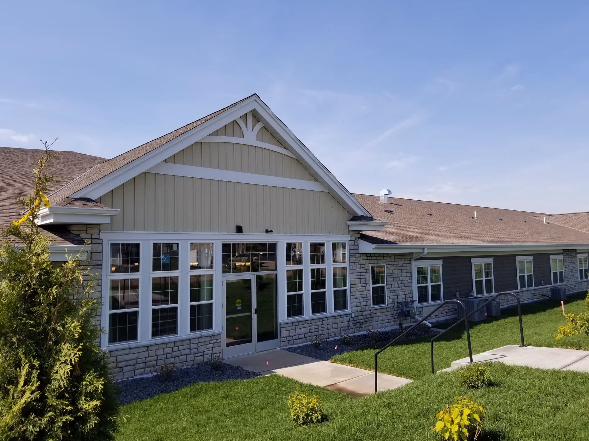 Exterior view of a single-story senior living facility building with beige siding, stone accents, multiple windows, and a glass door entrance. The building is surrounded by green grass and small shrubs under a clear blue sky.