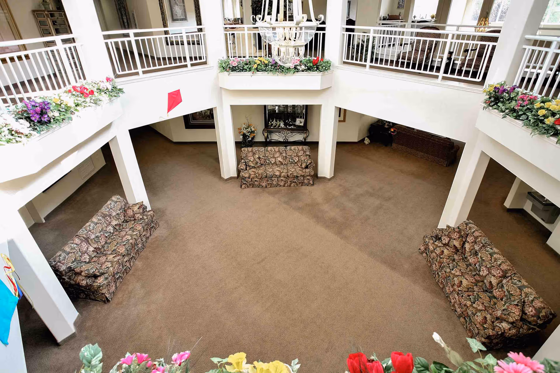 Spacious two-story common area in a senior living facility with floral-patterned sofas arranged around the perimeter. The upper level has white railings and flower boxes with colorful artificial flowers. A large chandelier hangs from the ceiling, and there are decorative items and furniture visible on the upper floor.