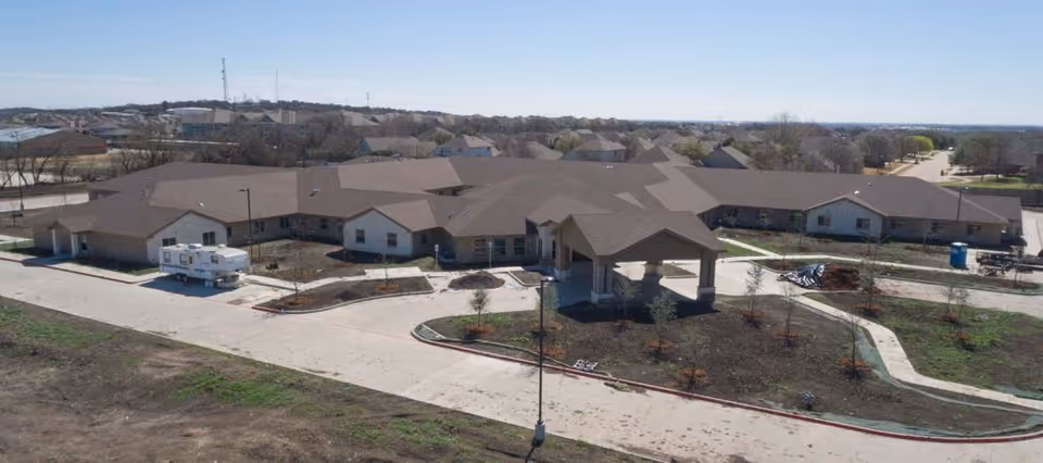 Aerial view of a single-story tan healthcare and rehabilitation building with multiple wings, a covered entrance drive, and surrounding landscaping and roads.