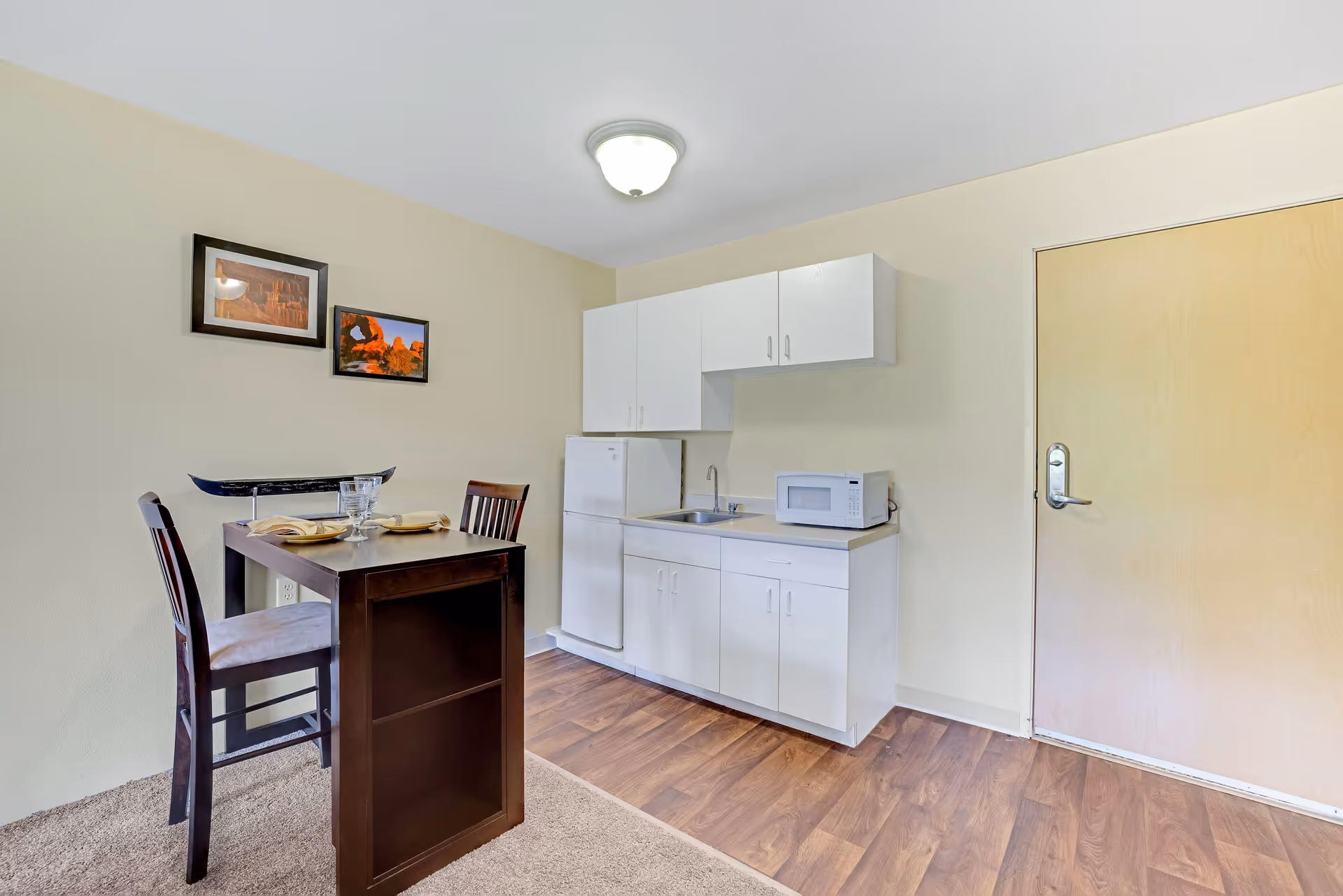 Small kitchenette area with white cabinets, a mini refrigerator, a microwave, and a sink. Next to the kitchenette is a small dark wooden dining table set for two with two chairs. Two framed pictures hang on the beige wall above the dining table. The floor is a combination of carpet and wood laminate, and there is a closed wooden door to the right.