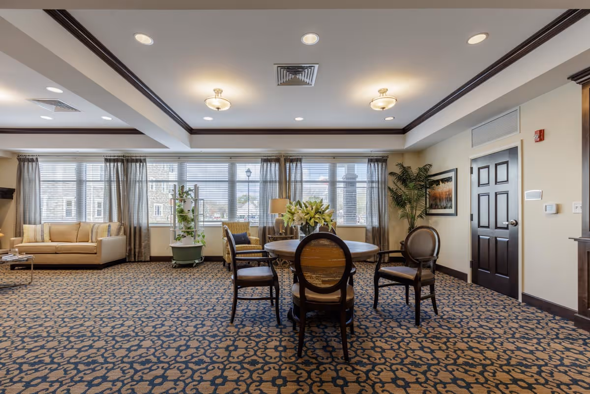 A spacious, well-lit common area with a patterned carpet, a round wooden table surrounded by four chairs, a beige sofa, an armchair, a tall plant, and large windows with sheer curtains allowing natural light to fill the room.