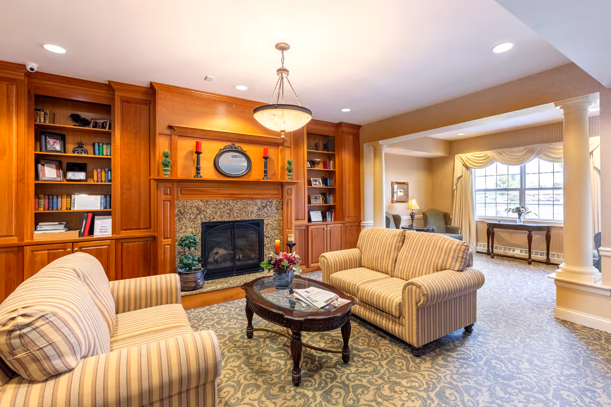 A cozy living room featuring striped sofas, a central coffee table, a fireplace surrounded by built-in wooden bookshelves, and a large windowed seating area.