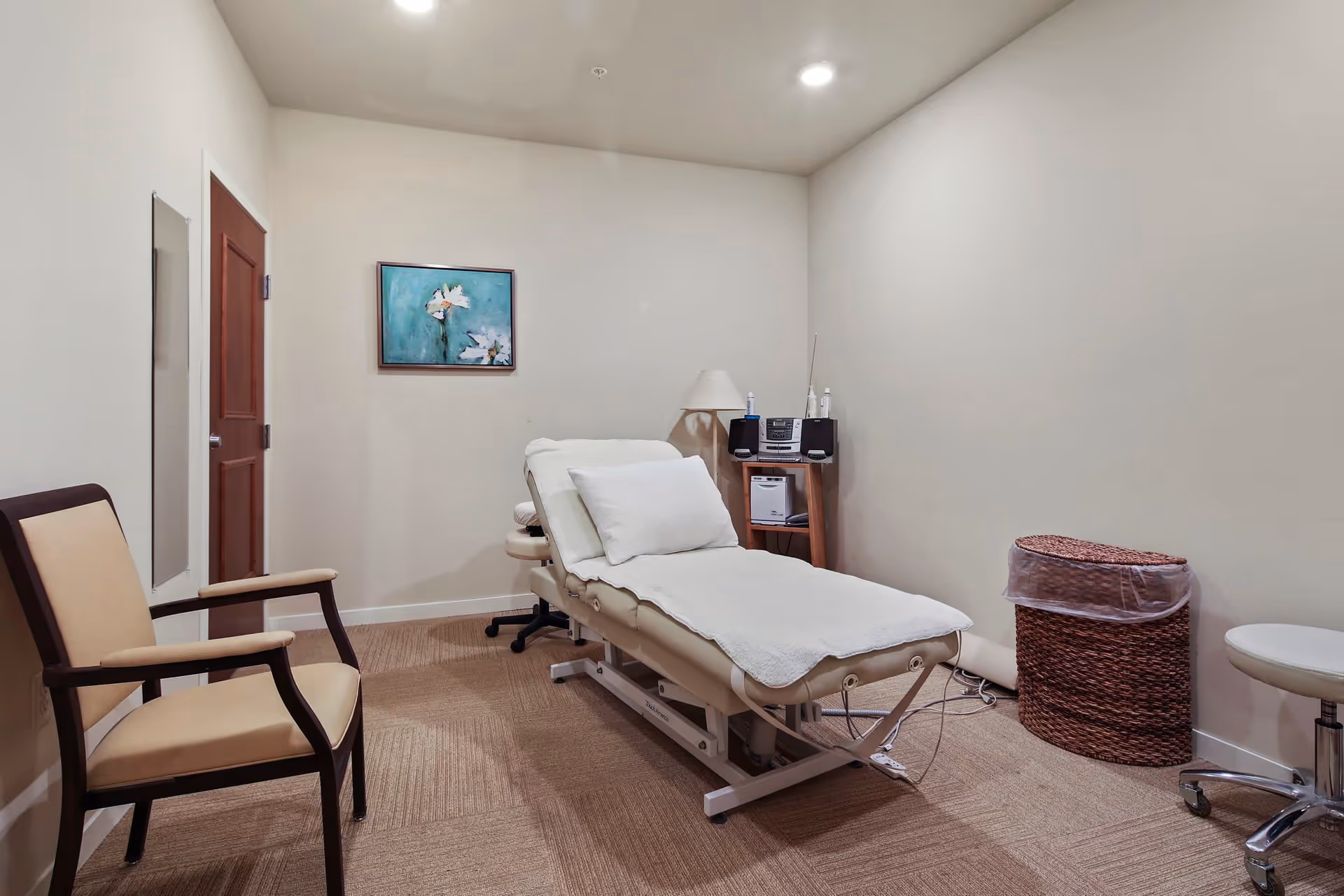 A small treatment room with a padded medical examination table covered with a white sheet and pillow. There is a beige armchair to the left, a small wooden table with a lamp and stereo system in the corner, a wicker trash bin with a plastic liner, and a white rolling stool. The walls are plain and light-colored, with a framed painting of white flowers on a blue background hanging above the examination table.