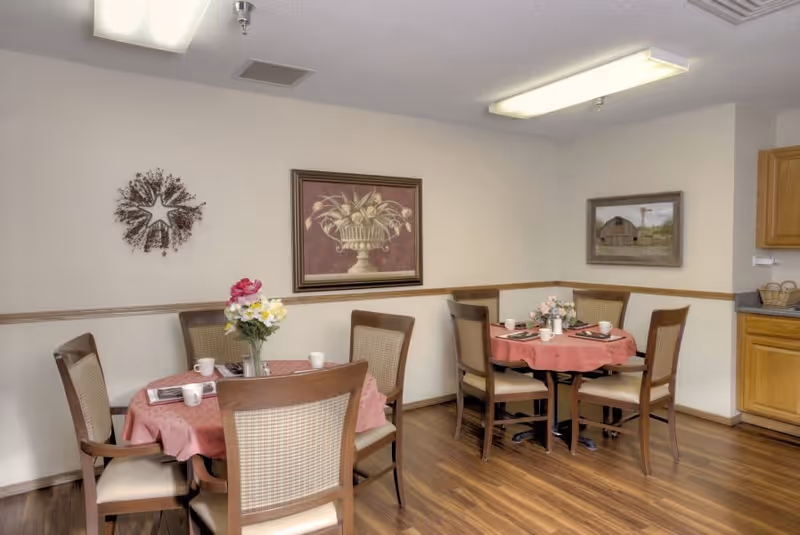 A dining area with two round tables covered with pink tablecloths, each surrounded by four wooden chairs with cushioned seats and backs. Each table has a floral centerpiece and place settings with cups and napkins. The room has wood flooring, light-colored walls with a wooden chair rail, and two framed paintings on the walls. There is a kitchen counter with wooden cabinets visible on the right side.