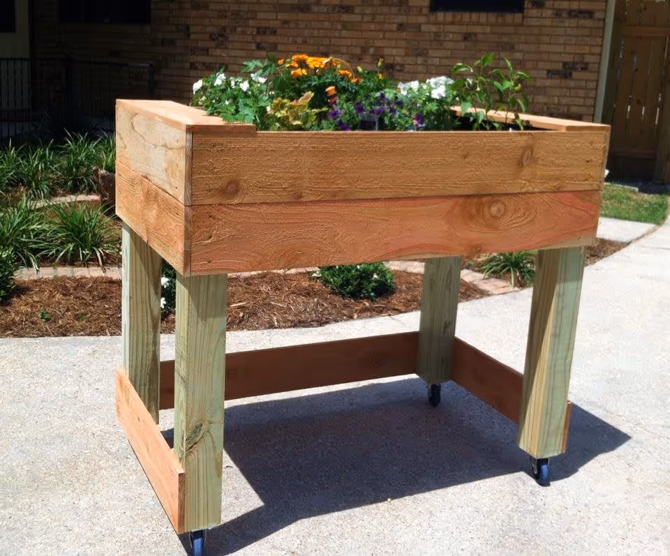 A raised wooden garden bed on wheels filled with various blooming flowers, placed outdoors on a concrete pathway with grass and plants in the background.