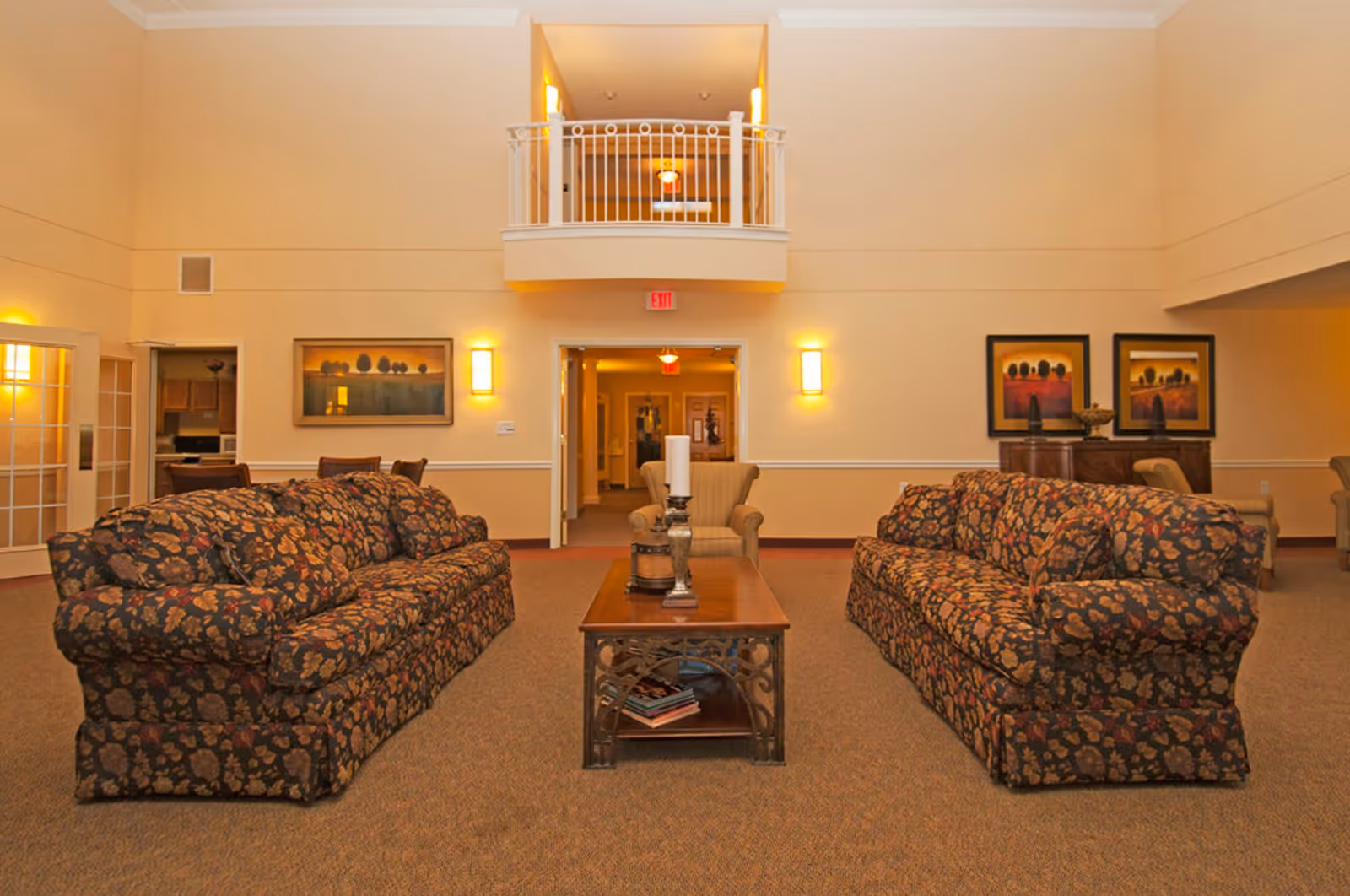 A spacious living room area with two floral patterned sofas facing each other and a wooden coffee table with decorative items in between. The room has beige walls, carpeted floor, wall-mounted lights, and framed artwork. There is a balcony railing above a doorway leading to a hallway, and additional seating and furniture are visible in the background.