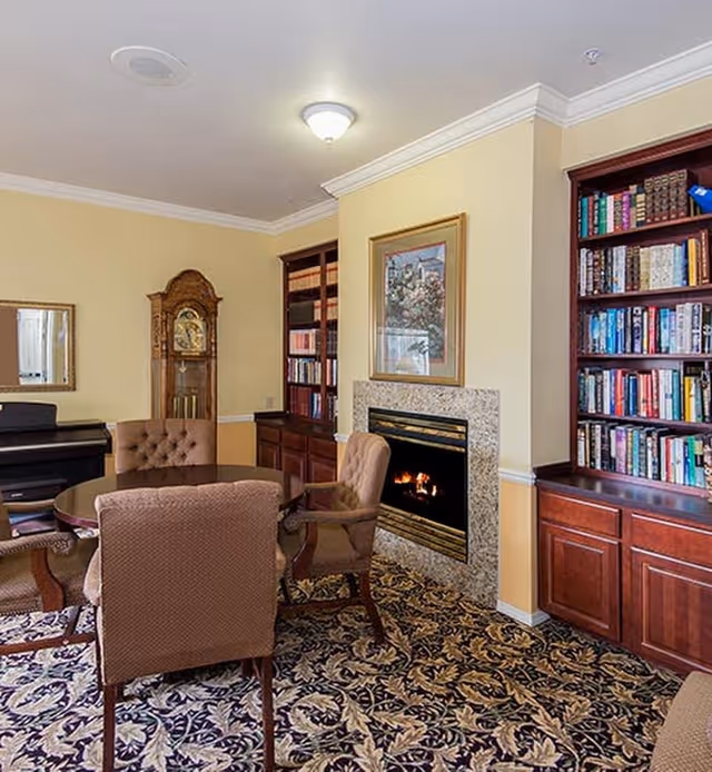 A cozy living room area with a round wooden table surrounded by four upholstered chairs. There is a fireplace with a granite surround and a framed painting above it. On either side of the fireplace are built-in wooden bookshelves filled with books. A grandfather clock and a piano are visible in the background. The room has beige walls and a patterned carpet.