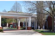 Exterior view of a senior living facility entrance with a covered drop-off area supported by white columns and brick bases, surrounded by trees and a driveway.