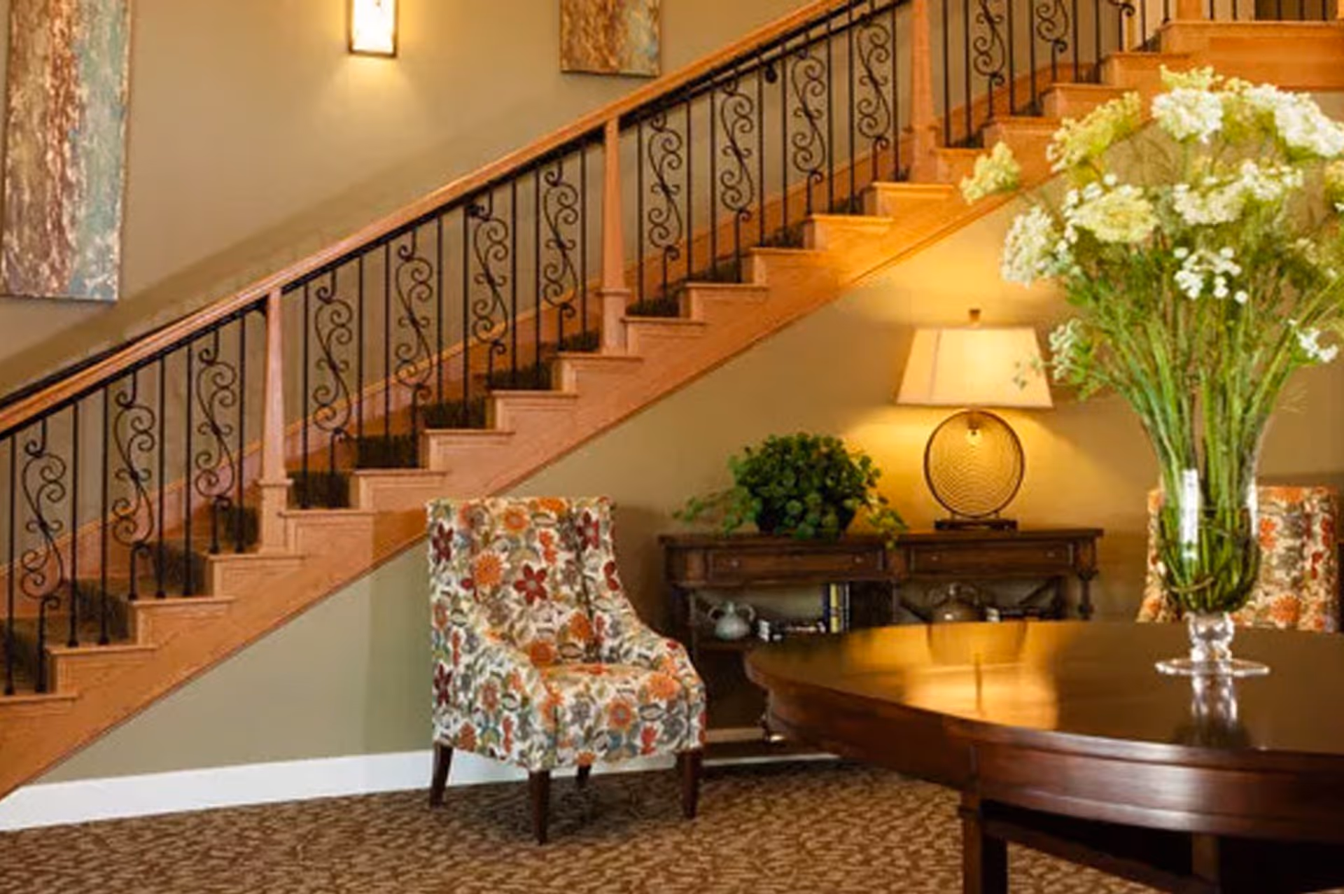 Interior view of a living area with a wooden staircase featuring decorative black iron railings. Below the staircase is a floral-patterned armchair, a wooden console table with a lamp and a plant, and a round wooden table with a large vase of white flowers.