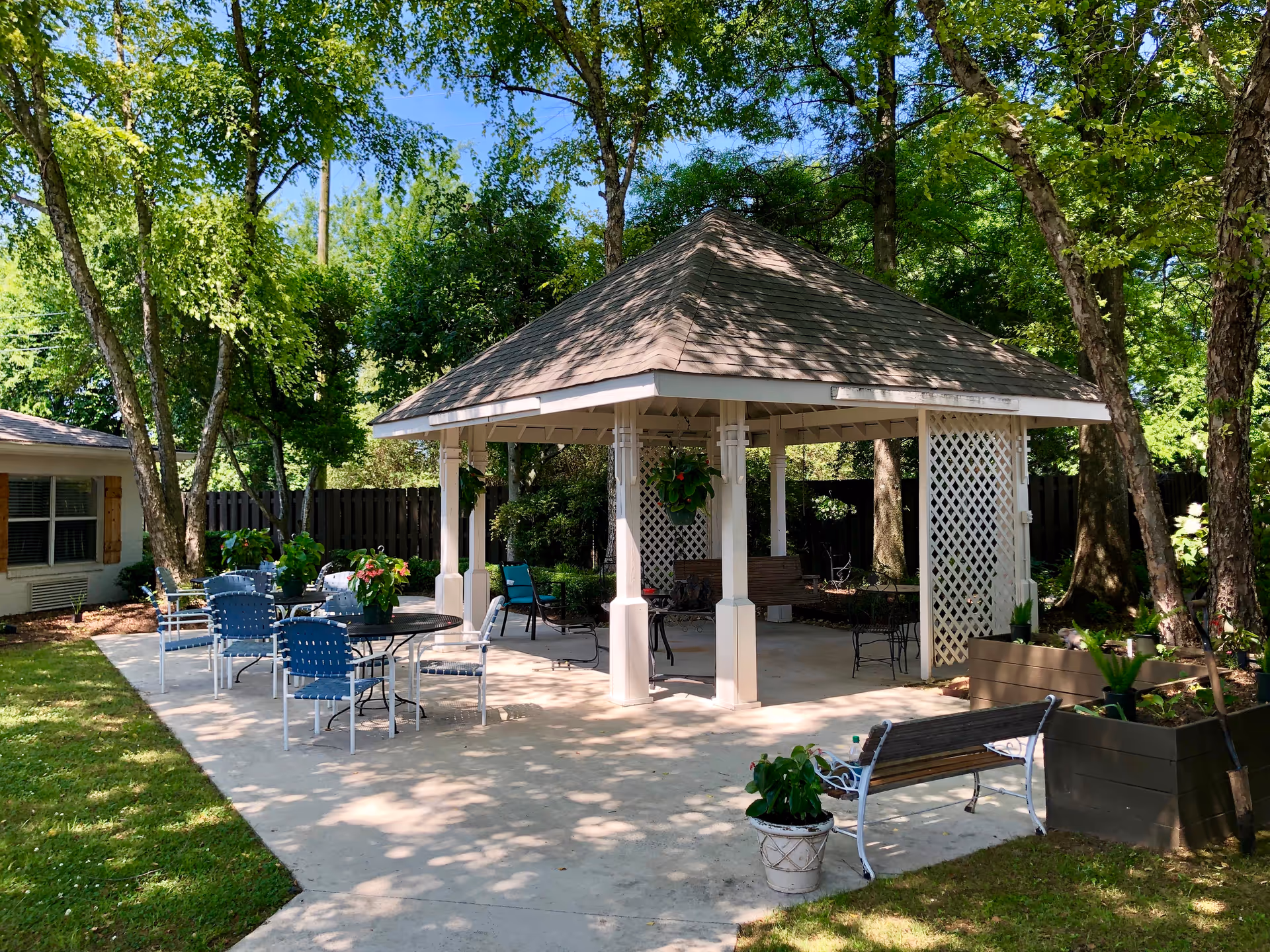 A white gazebo with tables, chairs, and benches on a shaded patio surrounded by trees and planters.