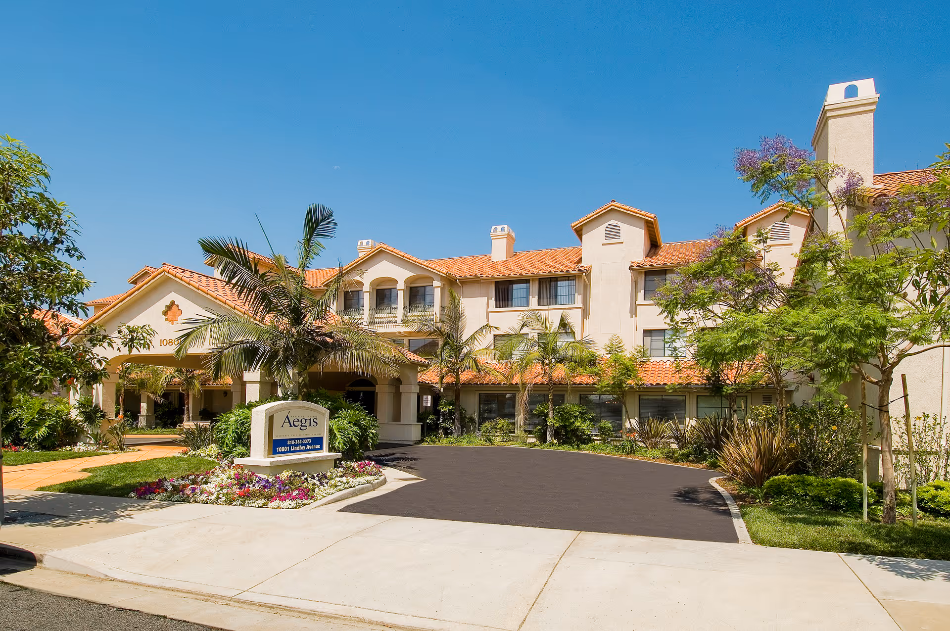 Exterior view of Aegis Living Granada Hills facility with a beige stucco building featuring red tile roofing, surrounded by palm trees and other greenery under a clear blue sky. A sign in front displays the facility name and address.