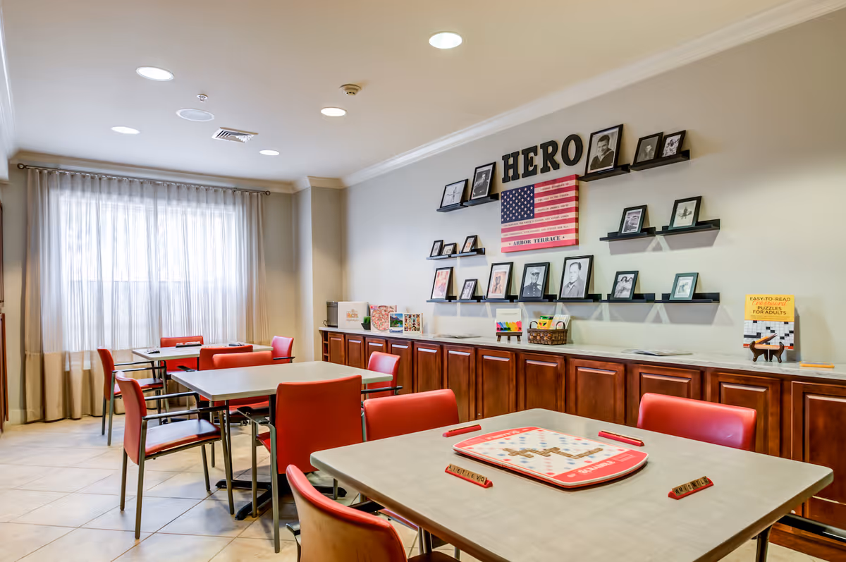 A bright room with several tables and red chairs arranged for playing board games. On one table, a Scrabble game is set up. The back wall features wooden cabinets topped with a marble surface, above which are shelves displaying framed photos and an American flag with the word 'HERO' above it. A large window with sheer curtains lets in natural light.