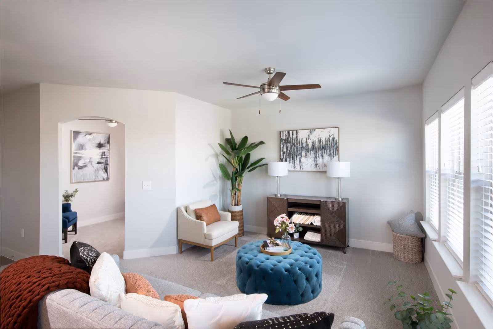 Bright modern living room with a sofa, armchair, tufted blue ottoman, console table, plants, and large windows.