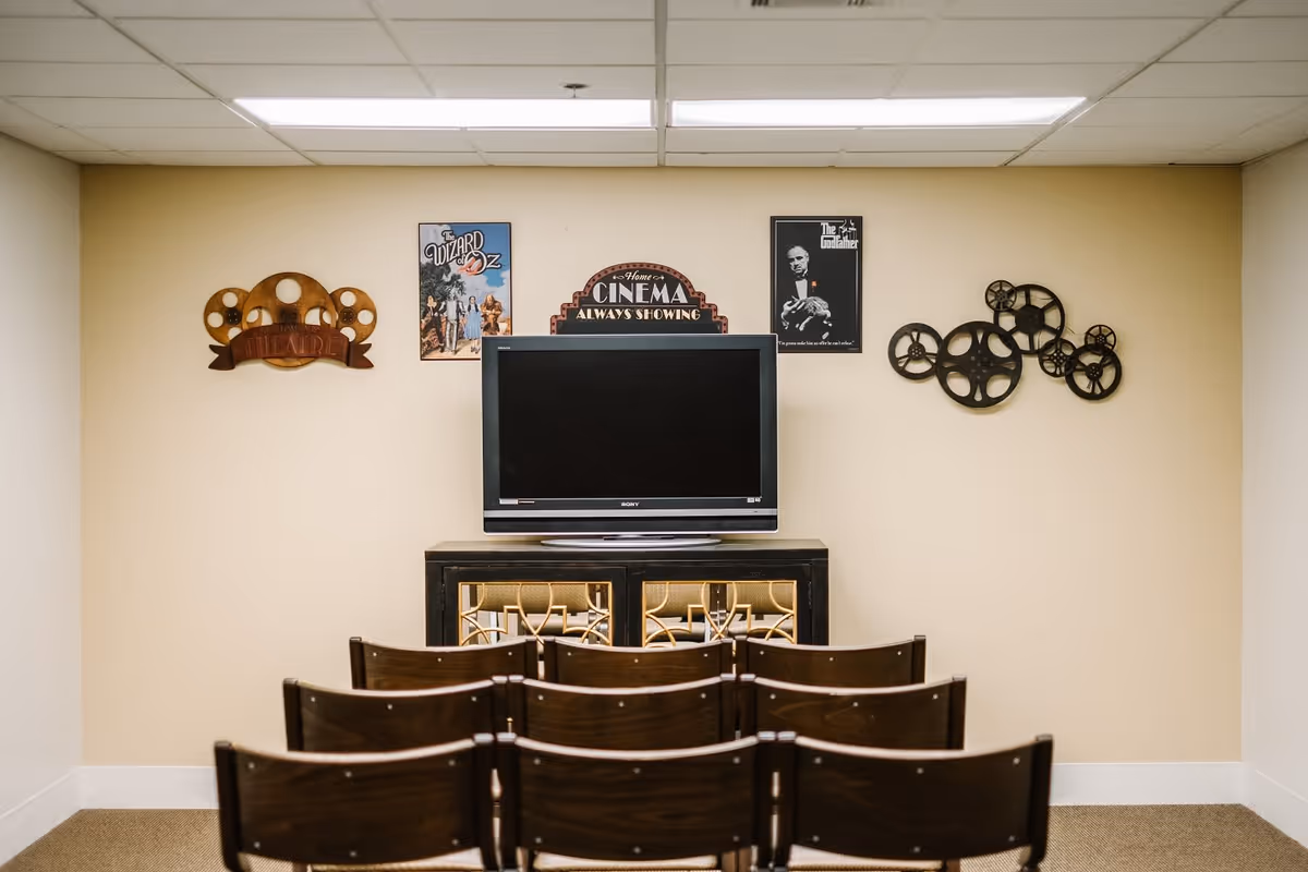 A small theater room with rows of wooden chairs facing a flat-screen TV mounted on a cabinet. The wall behind the TV is decorated with movie-themed art, including a sign that says 'Home Cinema Always Showing,' a poster of The Wizard of Oz, a poster of The Godfather, and decorative film reel wall art.