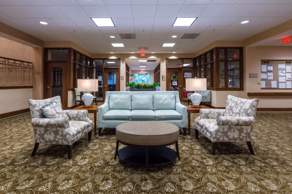 A communal seating area in a senior living facility lobby with a sofa, two patterned armchairs, table lamps, and a round ottoman.