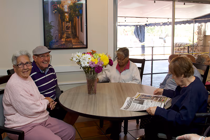 A group of elderly people sitting around a round wooden table in a well-lit room. There is a vase with colorful flowers in the center of the table. One person is reading a newspaper, and others are engaged in conversation or looking towards the camera. A large window with a view of an outdoor area is in the background, and a framed picture of a narrow street hangs on the wall.