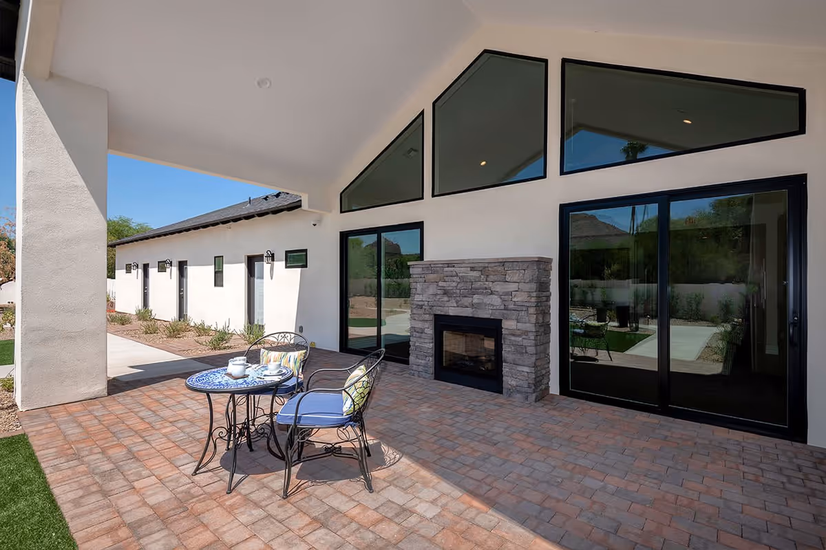 Covered outdoor patio with brick pavers, a small table and chairs, and a stone fireplace set against sliding glass doors.