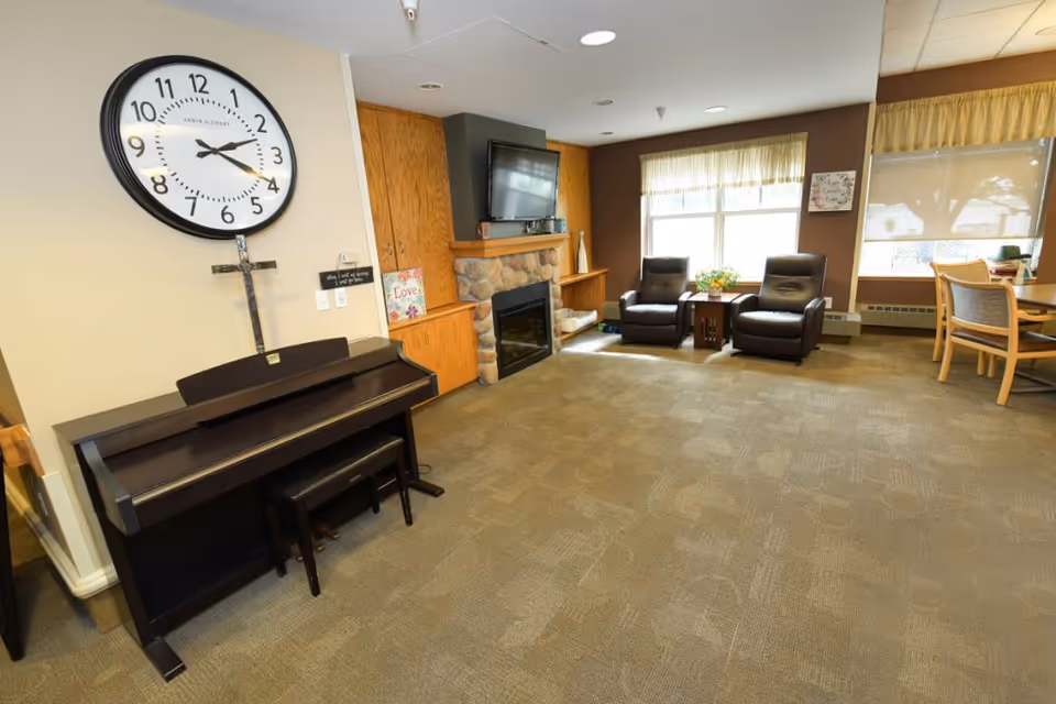 A cozy senior living common area featuring a large wall clock above a dark wooden piano with a bench. To the right, there is a stone fireplace with a mounted flat-screen TV above it, flanked by wooden cabinets. Two dark leather armchairs are positioned near a window with sheer curtains, next to a small wooden table with a flower arrangement. A dining table with chairs is visible in the background near another window with a yellow valance.
