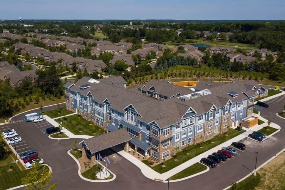 Aerial view of a large, modern senior living facility with multiple connected buildings featuring gray roofs and blue-gray siding with stone accents. The facility is surrounded by parking lots with cars and landscaped green areas. In the background, there is a residential neighborhood with many houses and trees under a clear blue sky.