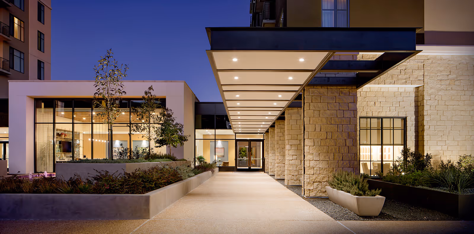 Evening view of the entrance to a modern senior living facility with a covered walkway, stone pillars, large windows, and landscaped planters on either side.