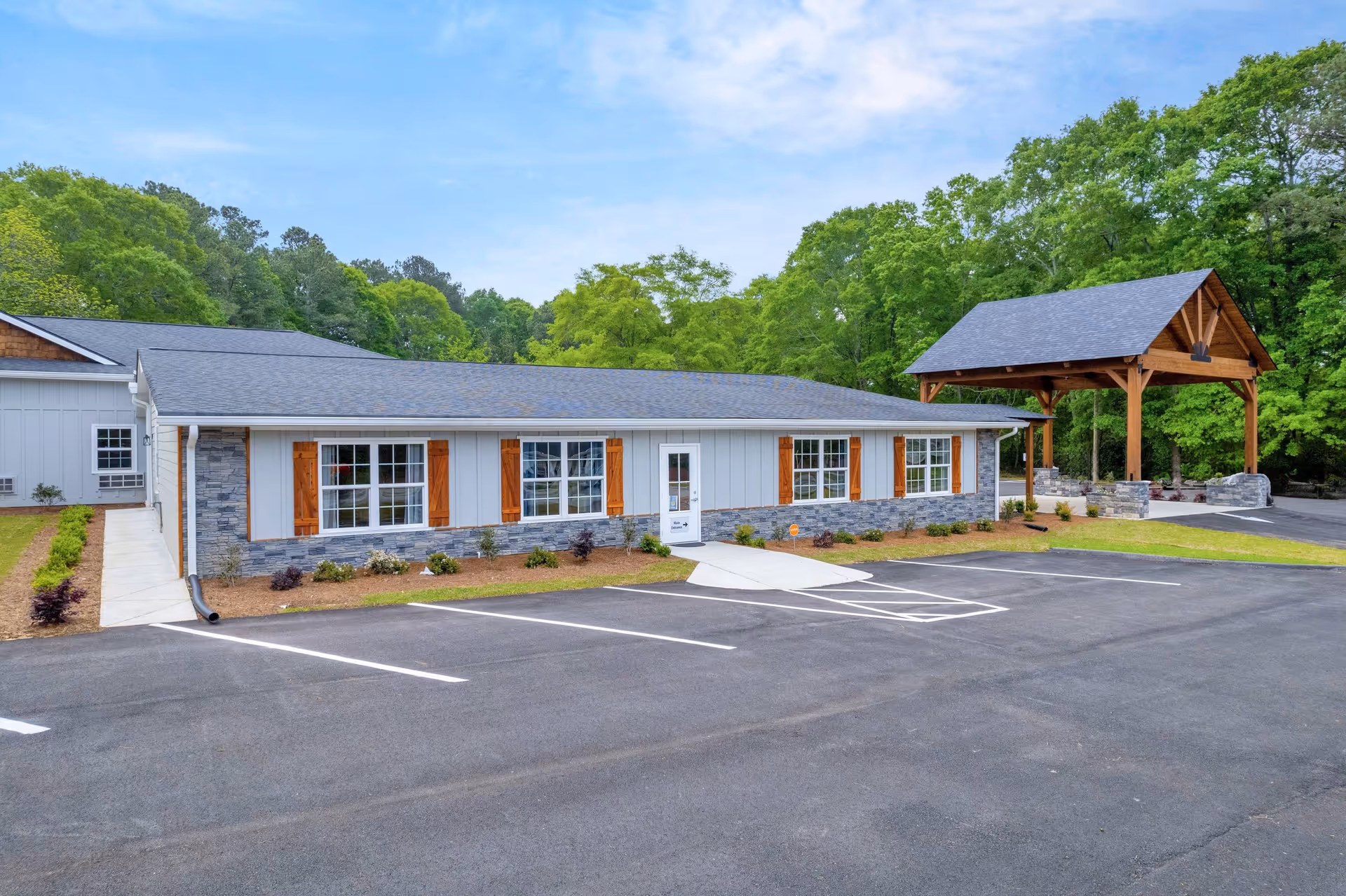 Exterior view of a single-story building with gray siding, stone accents, and wooden shutters on the windows. There is a small covered pavilion with wooden beams to the right, surrounded by green trees and a clear blue sky. The foreground shows an empty parking lot with marked spaces.