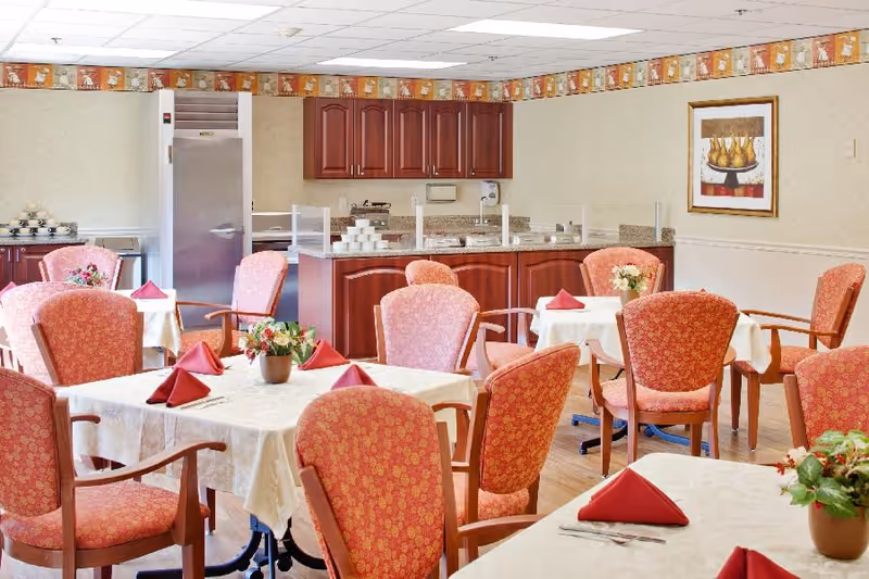 Bright dining room with several tables set with white tablecloths, red folded napkins, floral centerpieces, and upholstered chairs in front of a serving counter and refrigerator.