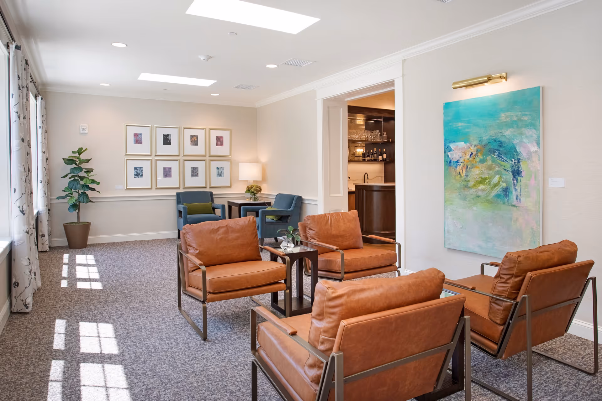 A bright and modern living room area with four brown leather armchairs arranged around a small wooden table. The room has large windows with patterned curtains allowing natural light to fill the space. On one wall, there is a large abstract painting in shades of green and blue, and on another wall, a series of framed pictures are displayed above two blue armchairs with green cushions. A potted plant and a table lamp add to the cozy ambiance. An open doorway reveals a bar area with shelves of glasses and bottles.