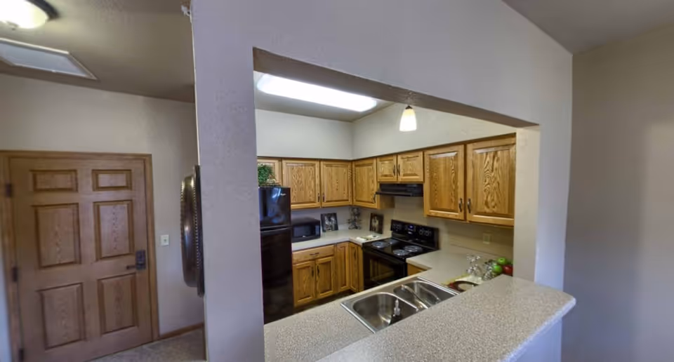 Small apartment-style kitchen with oak cabinets, black appliances, a double sink in a pass-through counter, and a wooden entry door visible.